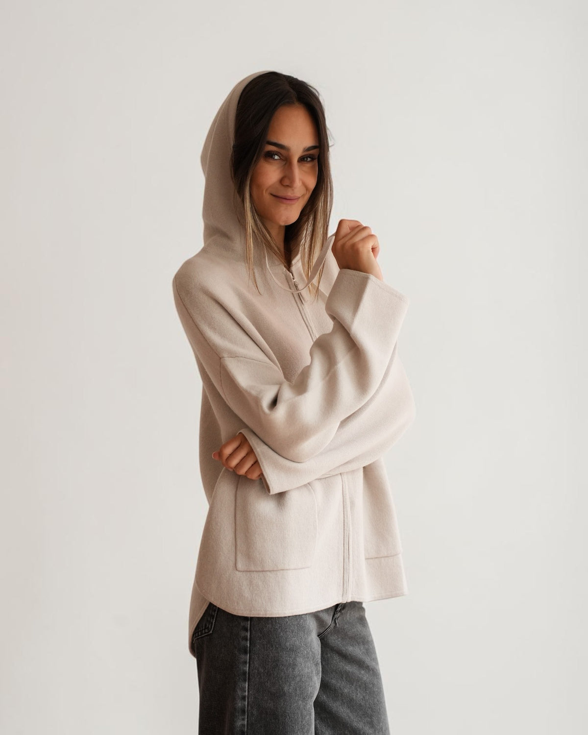 Hellgraue Kapuzen-Strickjacke am Model, Frontdetail mit Reißverschluss, aufgesetzten Taschen und weiten Ärmeln.