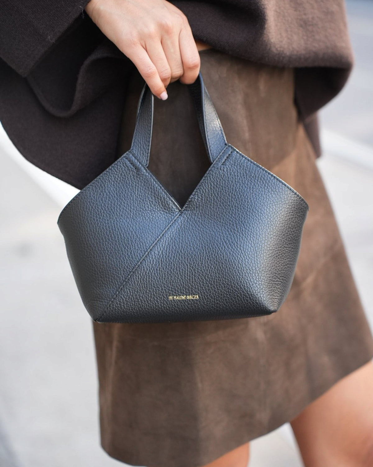 Close-up of a small black By Malene Birger leather bag with wide handles held by a model.